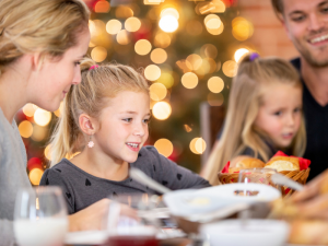 4 family members, 2 little girls and their parents, are eating holiday dinner with blurred holiday decorations in the background.